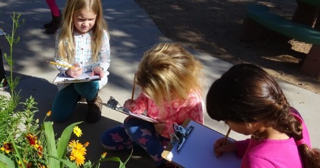 three preschool girls writing on a clipboard in a garden